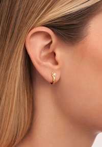 Orecchino a cerchio in oro con un design ondulato, caratterizzato da un piccolo accento di pietra preziosa trasparente. Texture liscia, finitura lucida e forma compatta.