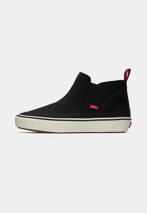 Black Vans slip-on sneaker with white sole, red logo tag on side, and red checkered pull tab on heel.