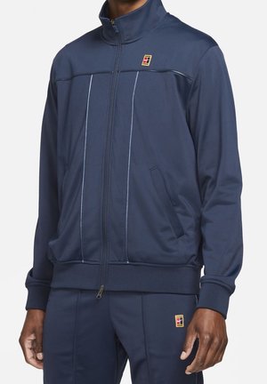 Homme portant une veste zippée bleu marine et un pantalon assorti avec de petits écussons logo rouges, jaunes et bleus sur la poitrine et la cuisse.