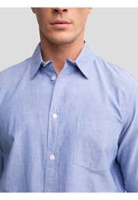 Bărbat purtând o cămașă bleu deschis cu nasturi și buzunar, gulerul ușor desfăcut, pe un fundal gri simplu.