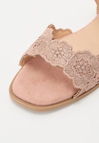 Rosa sandal med öppen tå och vågig blommig spetsrem samt en mjuk mocka sula på en ljusbrun sula.