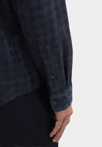 Donkerblauwe overhemdbloes met lange mouwen, voorzien van een tonale houndstooth-patroon, knoopmanchetten en een soepele stofstructuur. Getailleerde pasvorm zichtbaar.