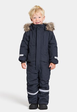 Combinaison de neige d'hiver bleu marine avec capuche doublée de fourrure, bandes réfléchissantes sur les manches et les jambes, fermeture éclair, et tissu doux au toucher. Bottes noires.