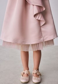 Kind trägt ein blassrosa Kleid mit gerüschtem Saum und passende rosa Schuhe mit Blumenriemen, steht auf einem hellen Boden.