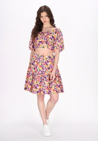 Blomstret crop top med puffærmer i gule, lilla og orange mønstre, parret med en matchende flared nederdel. Hvide sneakers fuldender looket.
