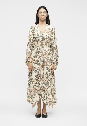 Γυναίκα που φοράει ένα κρεμ φόρεμα με λουλούδια, φαρδιά μανίκια και ζώνη, σε συνδυασμό με μαύρα επίπεδα παπούτσια, στέκεται μπροστά σε απλό φόντο.