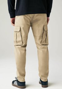 Pantaloni cargo bej cu multiple buzunare laterale, croi slim, realizați dintr-un material durabil, cu detalii vizibile de cusătură, purtați cu încălțăminte sport negre.