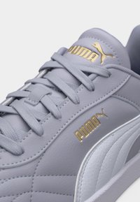 Ανοιχτό γκρι αθλητικό παπούτσι Puma με χρυσό λογότυπο Puma στη γλώσσα και στη πλευρά, υφασμένη λευκή ρίγα και κορδόνια σε ταιριαστό γκρι χρώμα.