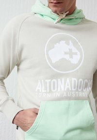 Sudadera en gris claro con capucha y bolsillo en verde menta. Presenta un logotipo gráfico blanco y el texto "ALTONADOCK NACIDO EN AUSTRALIA". Material de mezcla de algodón.