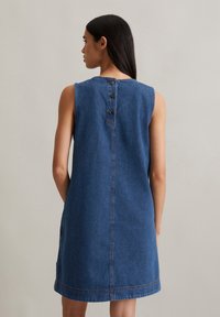 Denim jurk in middelblauw met een mouwloos ontwerp, voorzien van een ronde halslijn en drie houten knopen op de rug.