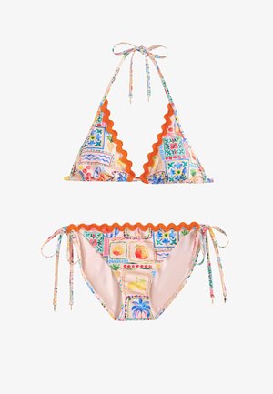 Mujer ajustándose los lazos de un bikini estampado y colorido en una playa soleada con palmeras y cielo azul de fondo.