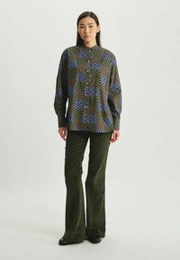 Chemise à boutons avec un motif géométrique bleu, vert et marron ; associée à un pantalon en velours côtelé olive à pattes d'éléphant et des chaussures noires.