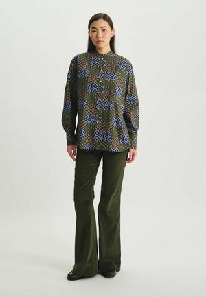 Chemise à boutons avec un motif géométrique bleu, vert et marron ; associée à un pantalon en velours côtelé olive à pattes d'éléphant et des chaussures noires.