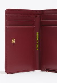 Interior de billetera de cuero color vino con múltiples ranuras para tarjetas, un cierre a presión dorado y un logo verde brillante en el lateral. Textura suave.