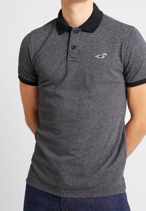 Hombre con una camiseta polo ajustada de color gris oscuro, con cuello negro y bordes de las mangas en negro, que presenta un pequeño logo de un pájaro blanco en el pecho.