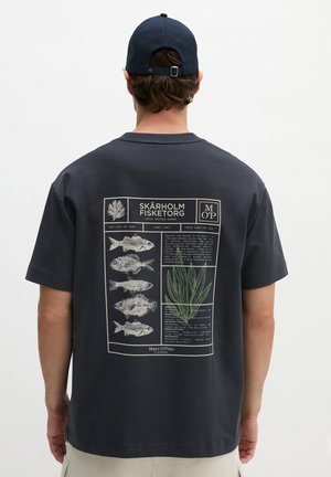 Mężczyzna w ciemnoszarej koszulce i granatowej czapce, tył koszulki przedstawia grafikę z pięcioma ilustracjami ryb i wodorostów oznaczonymi Skärholm Fisketorg.