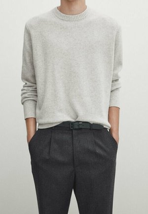 Person wearing a light gray crew neck sweater tucked into dark gray pleated trousers with a black belt, hands in pockets.