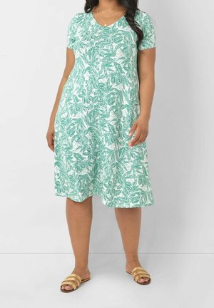 Vrouw die een knielange, korte mouwen, groen-witte jurk met bloemenmotief draagt, met gelaagde gouden halskettingen en gouden platte sandalen.