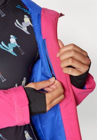 Slēpošanas jaka ar zilu virsmu un rozā akcentiem. Aprīkota ar rāvējslēdzēju, velcro aproci un flīsa oderi. Melna bāzes kārta ar slēpošanas grafiku.
