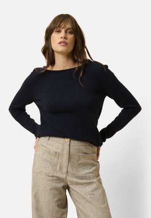Granatowy sweter dziany z szerokim dekoltem i długimi rękawami, zestawiony z jasnobeżowymi, teksturowanymi spodniami o wysokim stanie z kieszeniami.