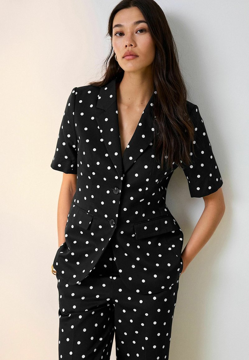 Femme aux longs cheveux foncés portant un blazer noir à manches courtes et un pantalon à pois blancs, debout contre un mur blanc.