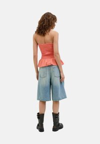 Peplum bluzica iz teksturiranega oranžnega blaga, v kombinaciji s svetlo modrimi denim kratkimi hlačami in črnimi vojaškimi škornji, prikazana s hrbtne strani.