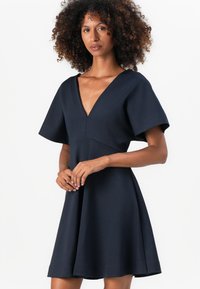 Vestido azul marino con mangas cortas acampanadas, escote en V y un cuerpo ajustado que se abre en una falda. Textura suave, diseño minimalista.