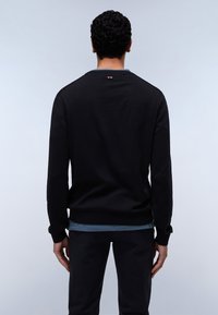 Uomo con capelli ricci che indossa una maglietta nera a maniche lunghe sopra una canotta blu, in piedi rivolto di spalle contro uno sfondo chiaro e uniforme.