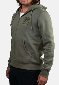 Homme portant un hoodie zippé vert olive avec des poches avant et un logo brodé "RS REDSKINS" sur la poitrine, associé à un pantalon noir.
