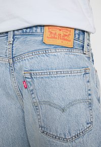 Jean en denim bleu clair avec une étiquette en cuir ornée de texte et de motifs embossés, poches arrière avec accents de couture, et une petite étiquette de marque.