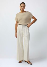 Beige gebreide top met korte mouwen, gecombineerd met crèmekleurige broek met wijde pijpen en een zwart leren riem. Zwarte haksandalen maken de look compleet.