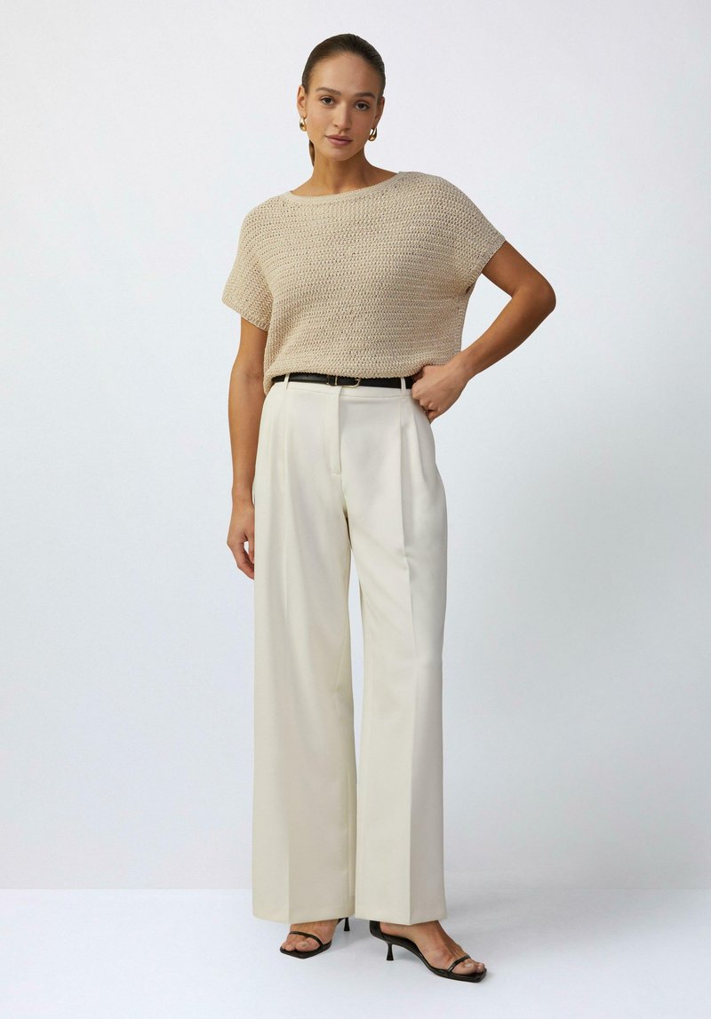 Beige gebreide top met korte mouwen, gecombineerd met crèmekleurige broek met wijde pijpen en een zwart leren riem. Zwarte haksandalen maken de look compleet.