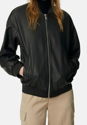 Frau trägt eine schwarze Leder-Bomberjacke mit Reißverschluss und gerippten Bündchen, kombiniert mit beigen Cargo-Hosen, die Hände in den Jackentaschen.