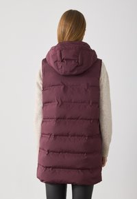 Langer kastanienbrauner Daunenweste mit Kapuze, die ein gesteppte Design und eine leicht glänzende Textur aufweist, getragen über einem hellbeigen Pullover.