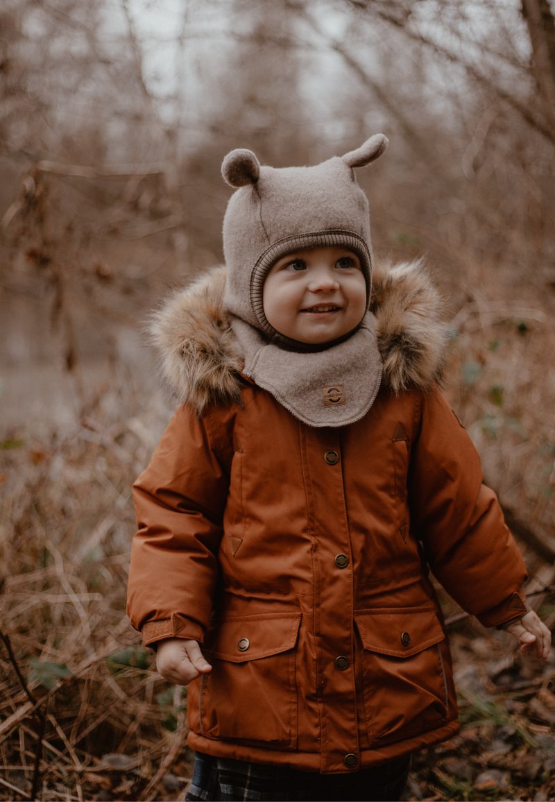 Hnedá nadýchaná bunda s kapucňou lemovanou kožušinou, vybavená dvoma prednými vreckami a zapínaním na gombíky. Béžová pletená čiapka s medvedími uškami. Textúrované pozadie.