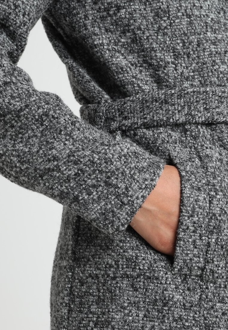 Cappotto di lana grigia testurizzata con maniche ampie, vita cinte e tasche laterali per un uso pratico. Il primo piano mette in risalto il dettaglio del tessuto.