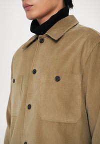 Smeđi kordura jakna s ovratnikom, koja ima dvije tamne gumbe na prsima i crne gumbe na prednjem zatvaranju.