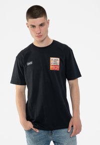 Black cotton t-shirt featuring a front patch with "Oasis" logo and graphic text. Short sleeves, crew neck, and relaxed fit.