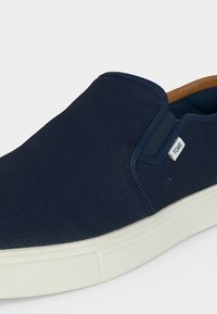 Baskets bleu marine sans lacets avec semelle blanche et petite étiquette blanche TOMS sur le côté, avec des panneaux latéraux élastiques et une doublure intérieure beige.