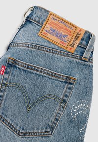 Jean en denim bleu clair avec étiquette en cuir marron, coutures jaunes et rivets décoratifs en argent sur la poche. Il présente un design classique à cinq poches.