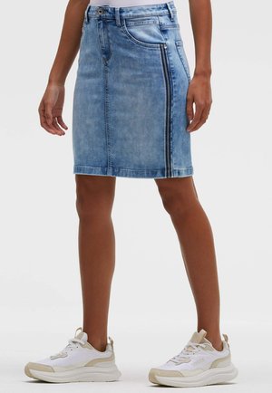 Knielanger hellblauer Jeansrock mit Reißverschlussdetail vorne, getragen mit weißen und beigen Sneakern bei einer stehenden Person.