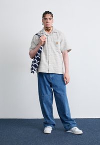 Kurzärmliges, hellgraues Shirt mit strukturierten Streifen und Logo. Weite blaue Jeans, weiße Sneaker und eine gemusterte Tasche in Marineblau und Weiß.