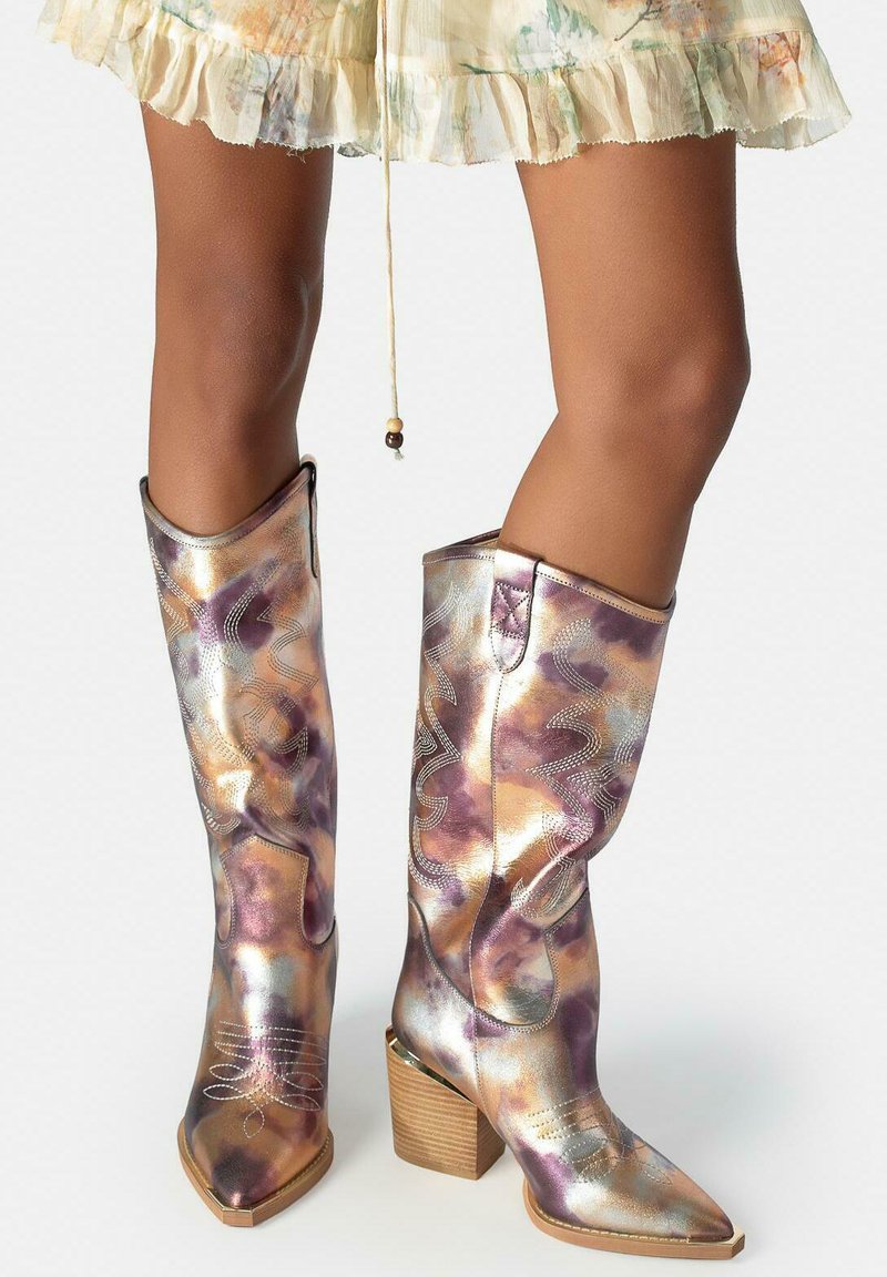 Metallic mehrfarbige Cowboy-Stiefel mit hohen Absätzen und aufgenähten Mustern, getragen zu einem leichten, blumigen Rüschrock.