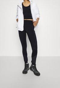 Czarne legginsy sportowe zestawione z czarnym topem crop i białą lekką kurtką. Legginsy mają ciemnoszare akcenty, a buty są czarne z odblaskowymi detalami.