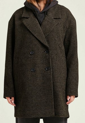Double-breasted dark brown and black houndstooth coat worn over a black hoodie, covering torso and arms, hands relaxed by sides.