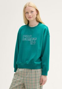 Turkusowy sweter z grafiką "COAST" w białym kolorze, okrągły dekolt, ściągacze przy rękawach i u dołu. W połączeniu z materiałowymi spodniami w kratę w neutralnych kolorach.