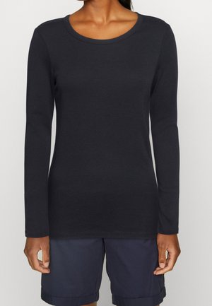 Personne portant un t-shirt noir uni à manches longues et un short bleu marine, debout devant un fond neutre.