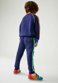 Completo sportivo blu navy con strisce arcobaleno sulle maniche e sui lati; dettaglio arcobaleno ricamato sui pantaloni; sneakers colorate con cinturini in velcro.