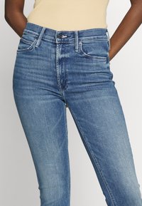 Jeans de denim en azul medio con un diseño ajustado, cintura alta, estilo de cinco bolsillos y una textura ligeramente desteñida. Cierre de botón y cremallera.