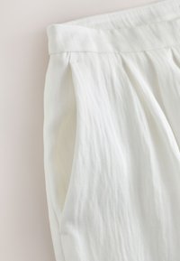 Pantalon blanc taille haute montrant la ceinture, des plis et une poche latérale avec une texture subtile du tissu et des détails de couture.